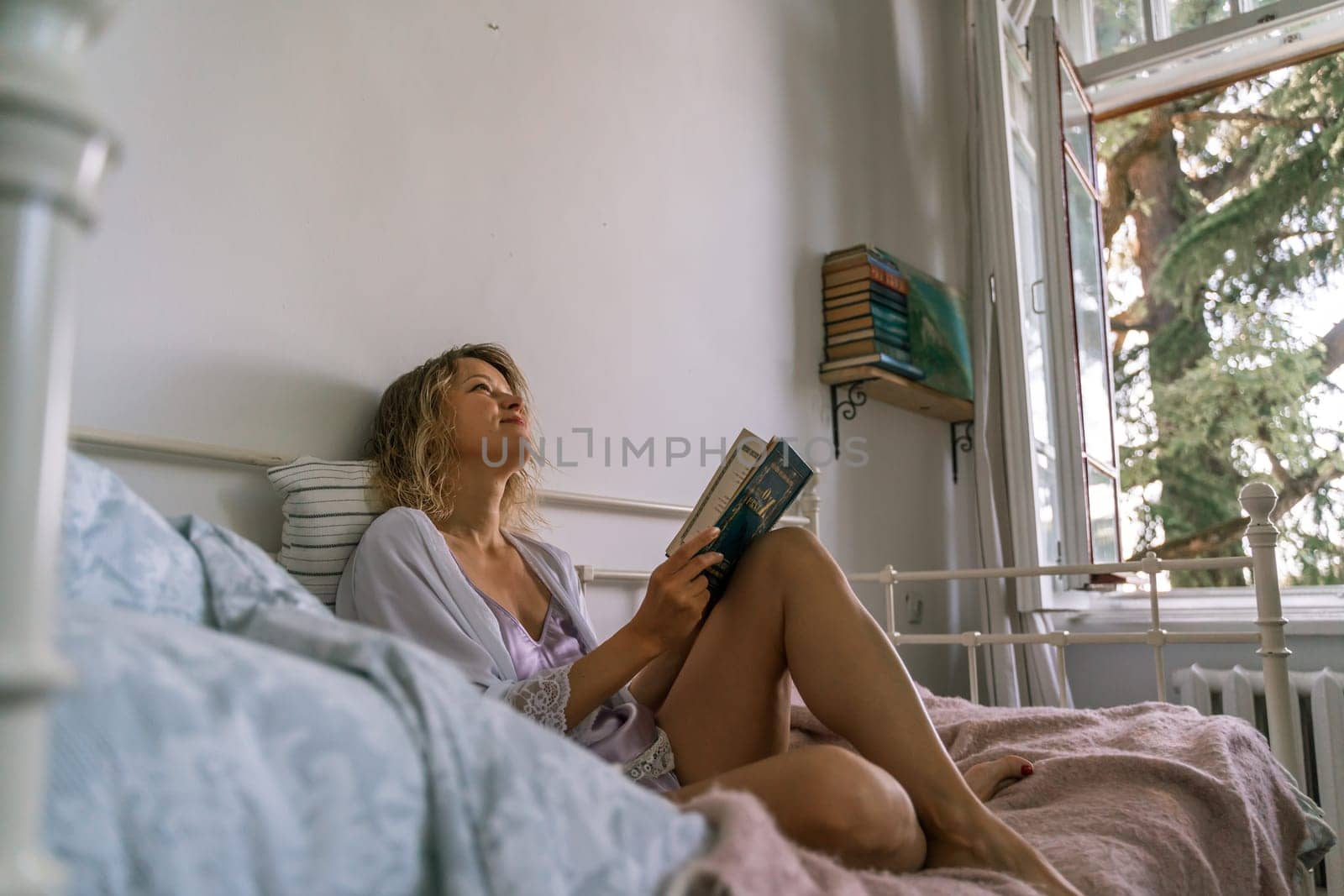 Woman reading a book in bed, lying on her stomach smiling happy and relaxed on a leisure day at home by Matiunina