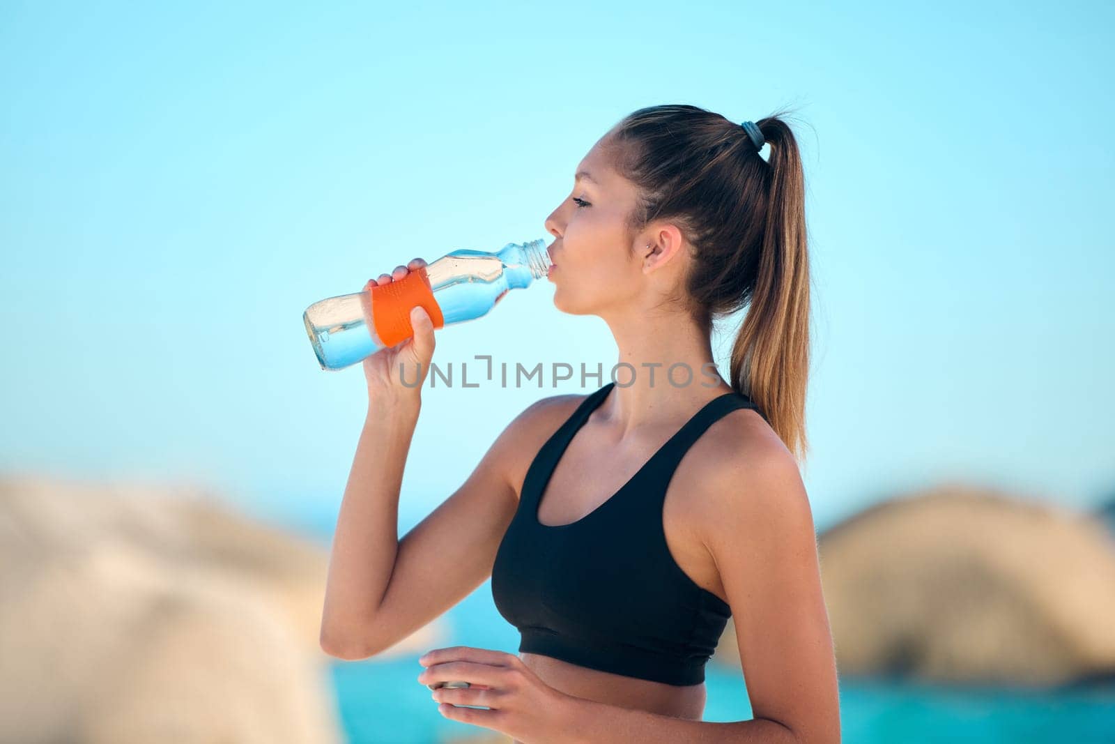 Beach, exercise and woman drinking water, health and training with stress relief, relax and break. Female person, girl and athlete outdoor, liquid and workout goal with hydration, fitness or wellness by YuriArcurs