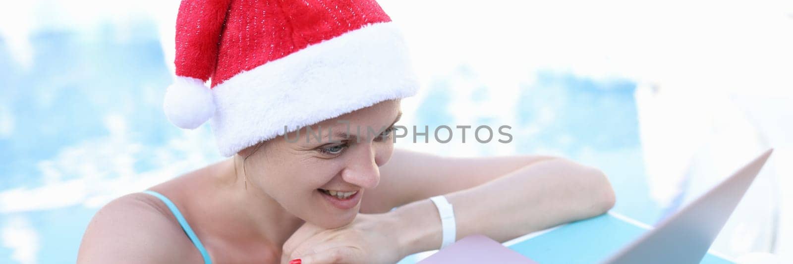 Woman in red santa claus hat looking at laptop on sunbed. Removed merry christmas and happy new year greetings concept