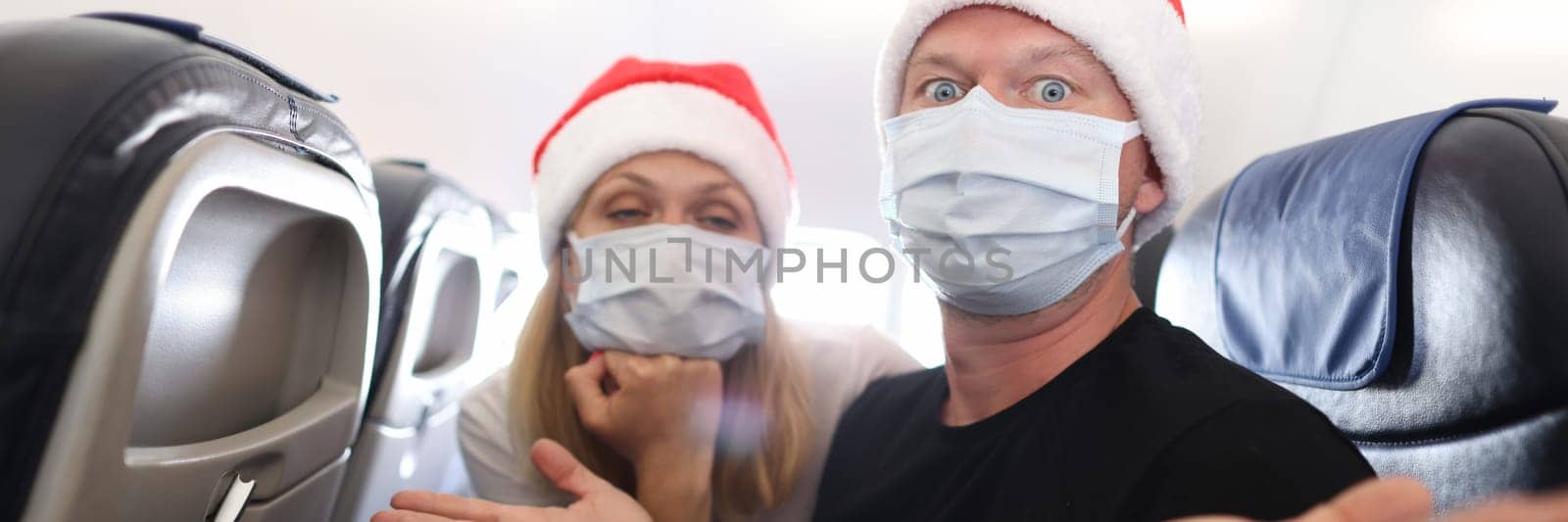Sad and drunk man and woman in protective medical masks and santa claus red caps flying in airplane. Difficulties of new year journey during covid19 pandemic concept