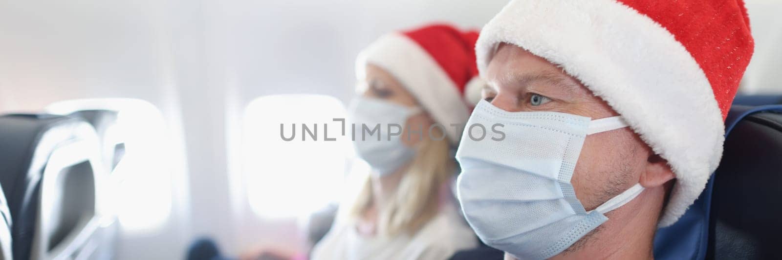 Man and woman in santa hats and protective face masks flying in airplane by kuprevich