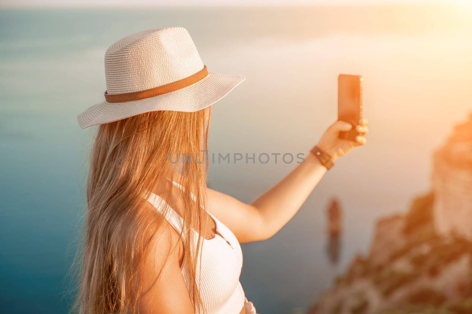 Selfie woman in a hat, white tank top, and shorts captures a selfie shot with her mobile phone against the backdrop of a serene beach and blue sea