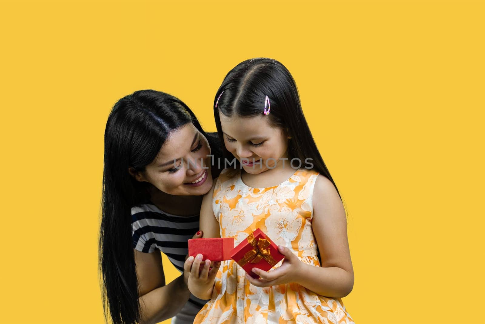 Little girl is opening a red gift box together with her mom. Isolated on yellow background.