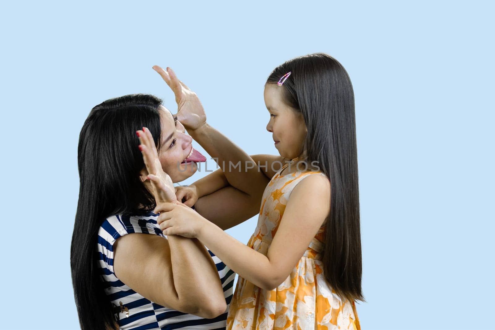 Young mother is making funny faces to her little daughter. Brunette woman amusing a little girl. Isolated on pale blue.