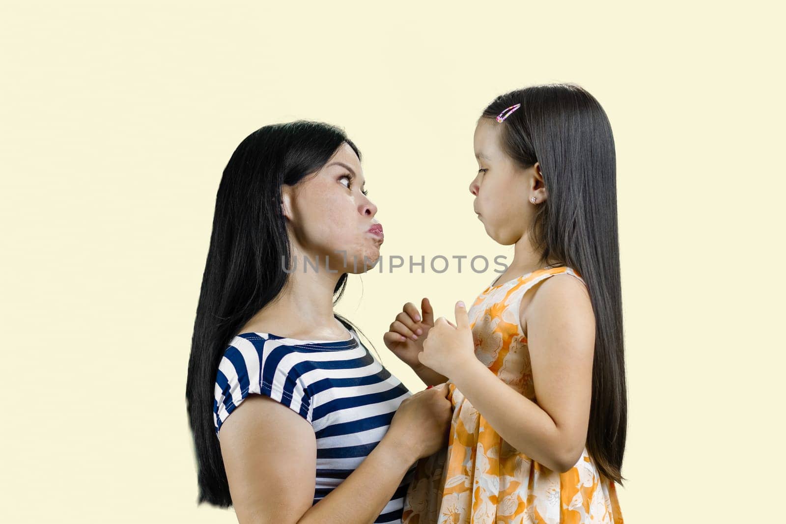 Side view of mother and her little girl are making faces to each other. Isolated on pastel yellow.