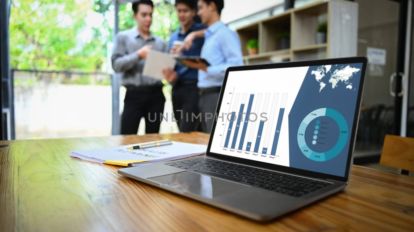 Laptop computer with financial graphs and statistics on monitor on wooden office desk and businesspeople standing in background.