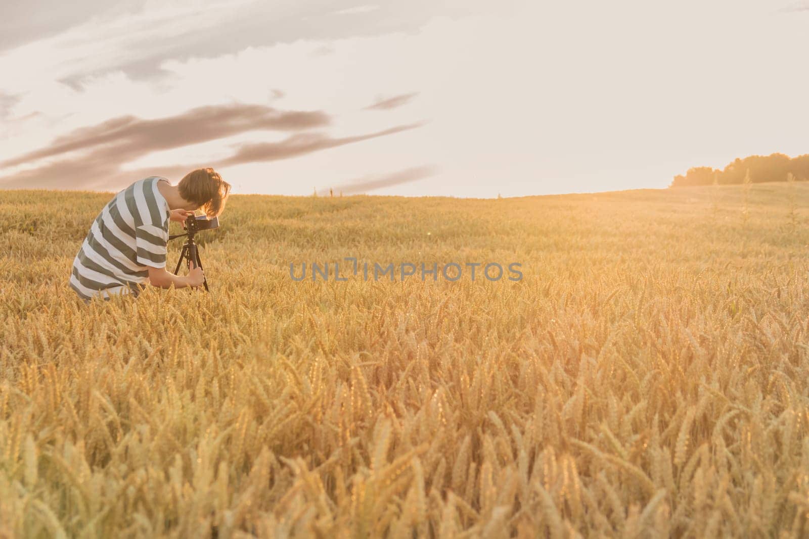 A young man in a striped T-shirt shoots a sunset in a wheat field on a phone that is mounted on a tripod..