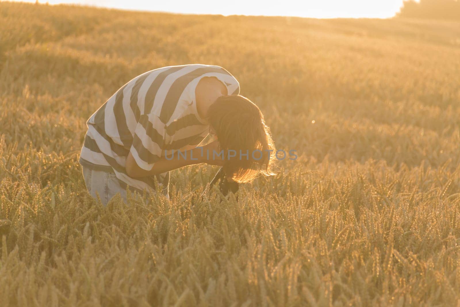 A young man in a striped T-shirt shoots a sunset in a wheat field on a phone that is mounted on a tripod. by Alla_Yurtayeva