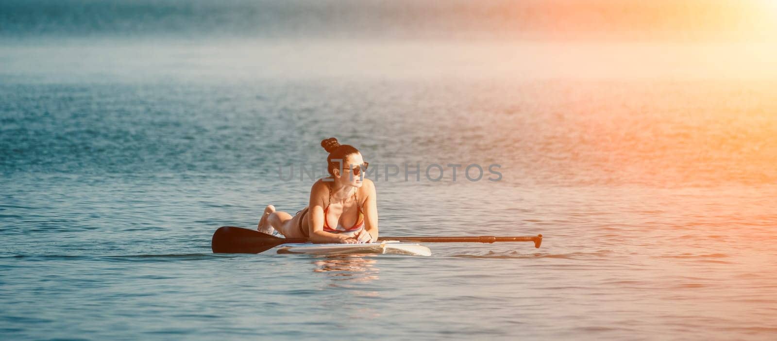 Silhouette of woman standing, surfing on SUP board, confident paddling through water surface. Idyllic sunset or sunrise. Sports active lifestyle at sea or river.