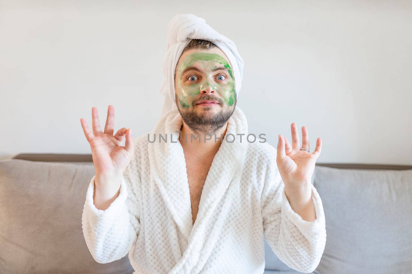 Funny man enjoys beauty treatments and luxury relaxation at home spa. A man with a towel on his head sits on a sofa in a white bathrobe and with a cosmetic mask on his face
