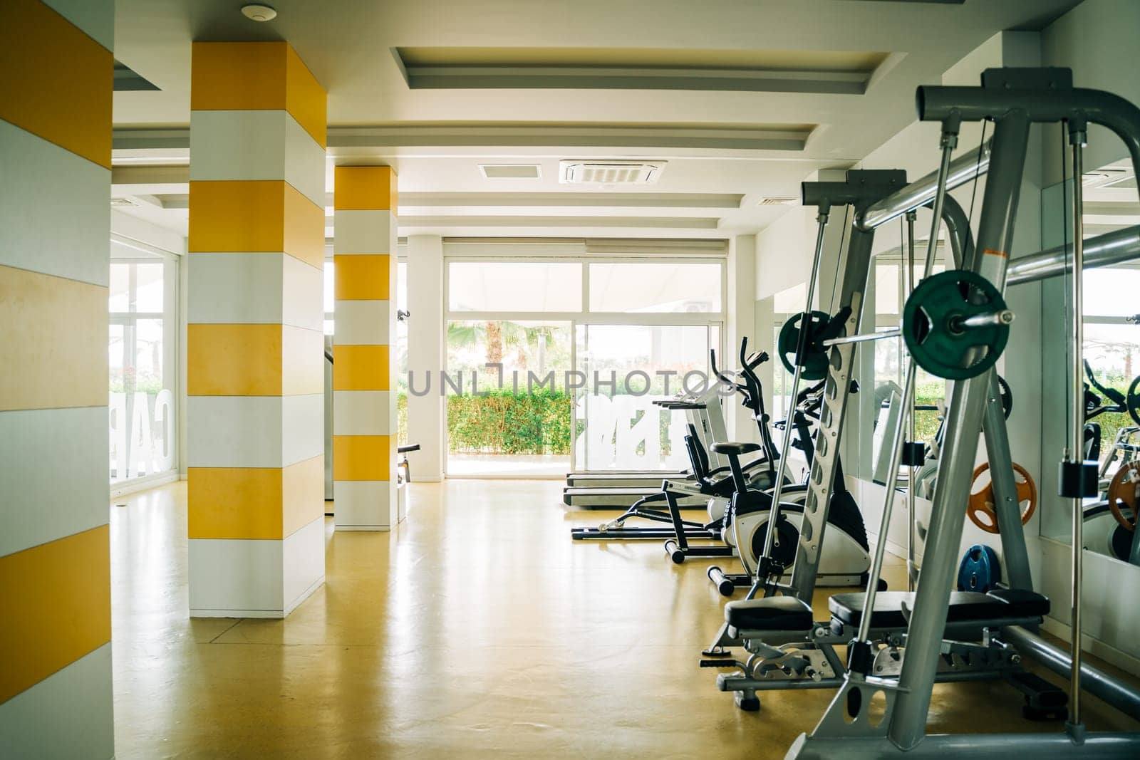 Gym Room Fitness Center With Various Equipment And Machines with views of palm and trees in Background of Large Glass Windows.