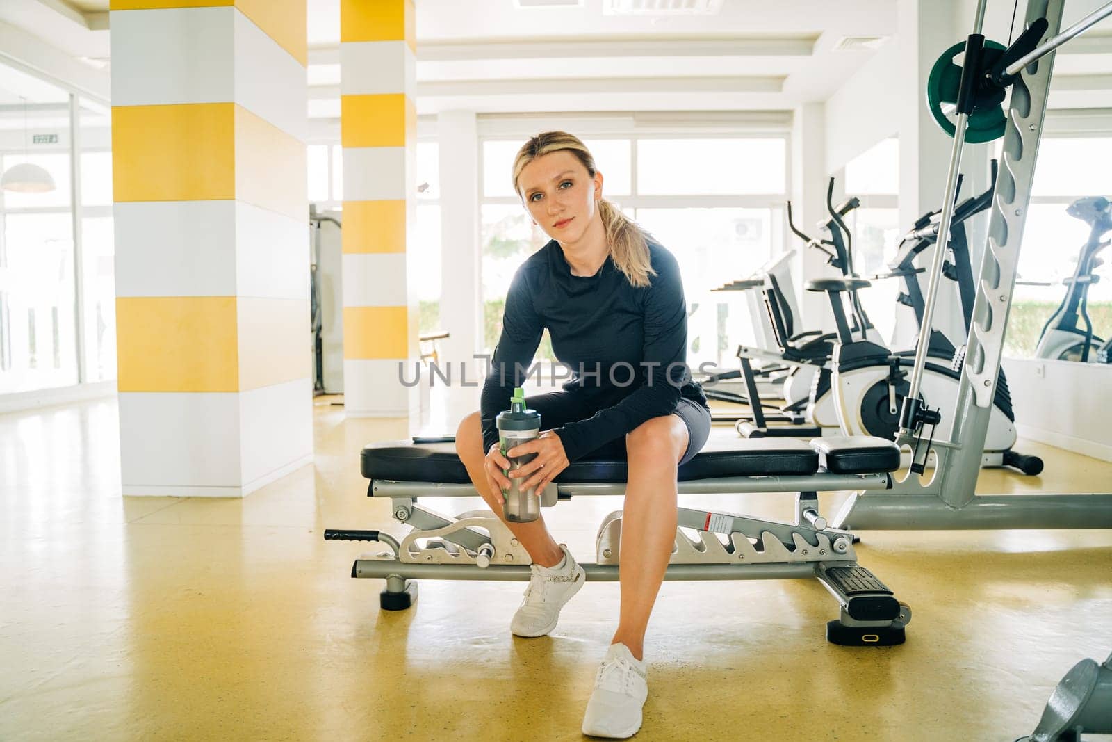 Relaxing after workout. Beautiful young woman sitting on bench at gym. Young sport woman at gym taking a break from training and drinking water