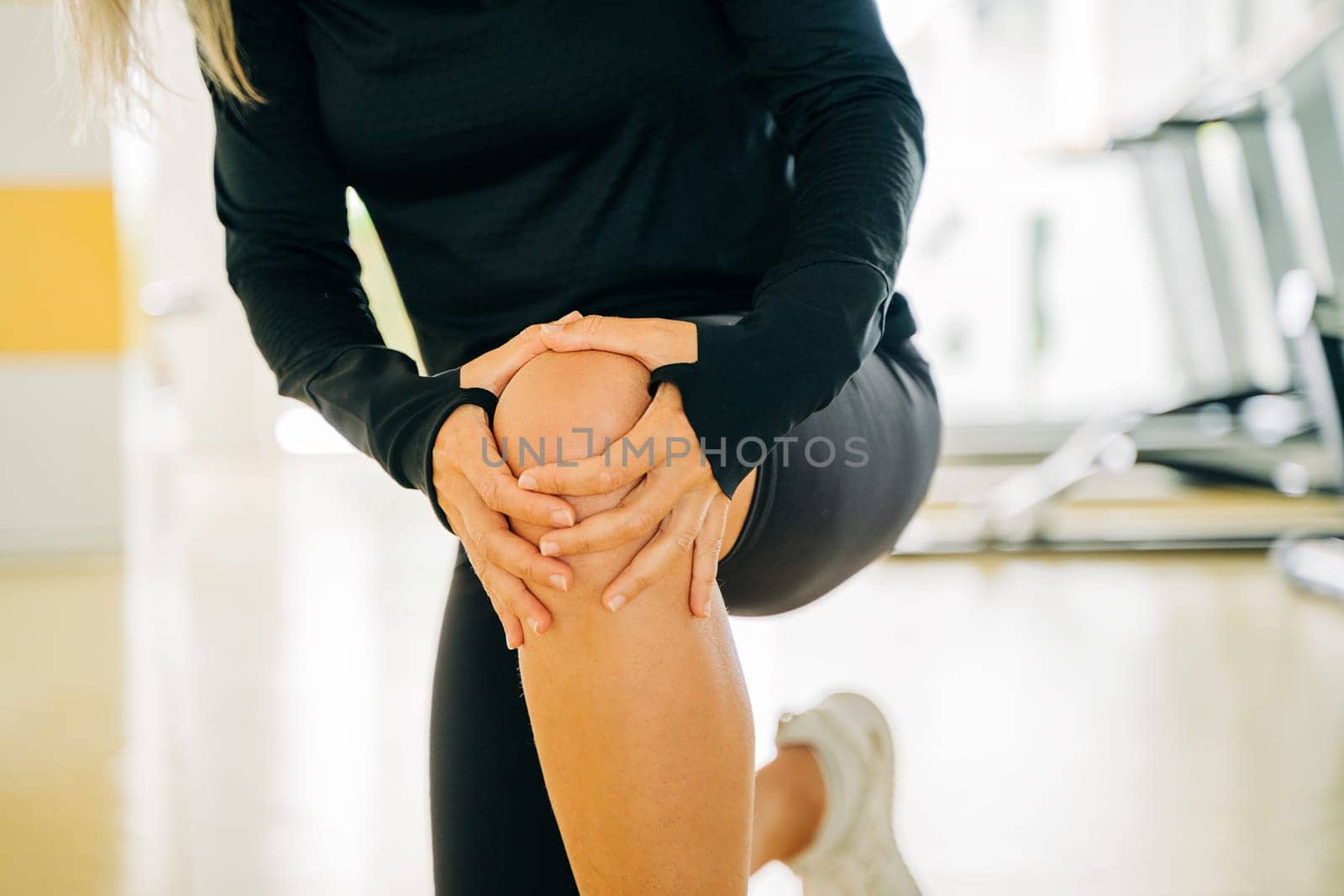 Close up woman holding knee with hands in pain after suffering injury gym workout. Fitness sport woman sit on bench with knee injury and pain by Ostanina