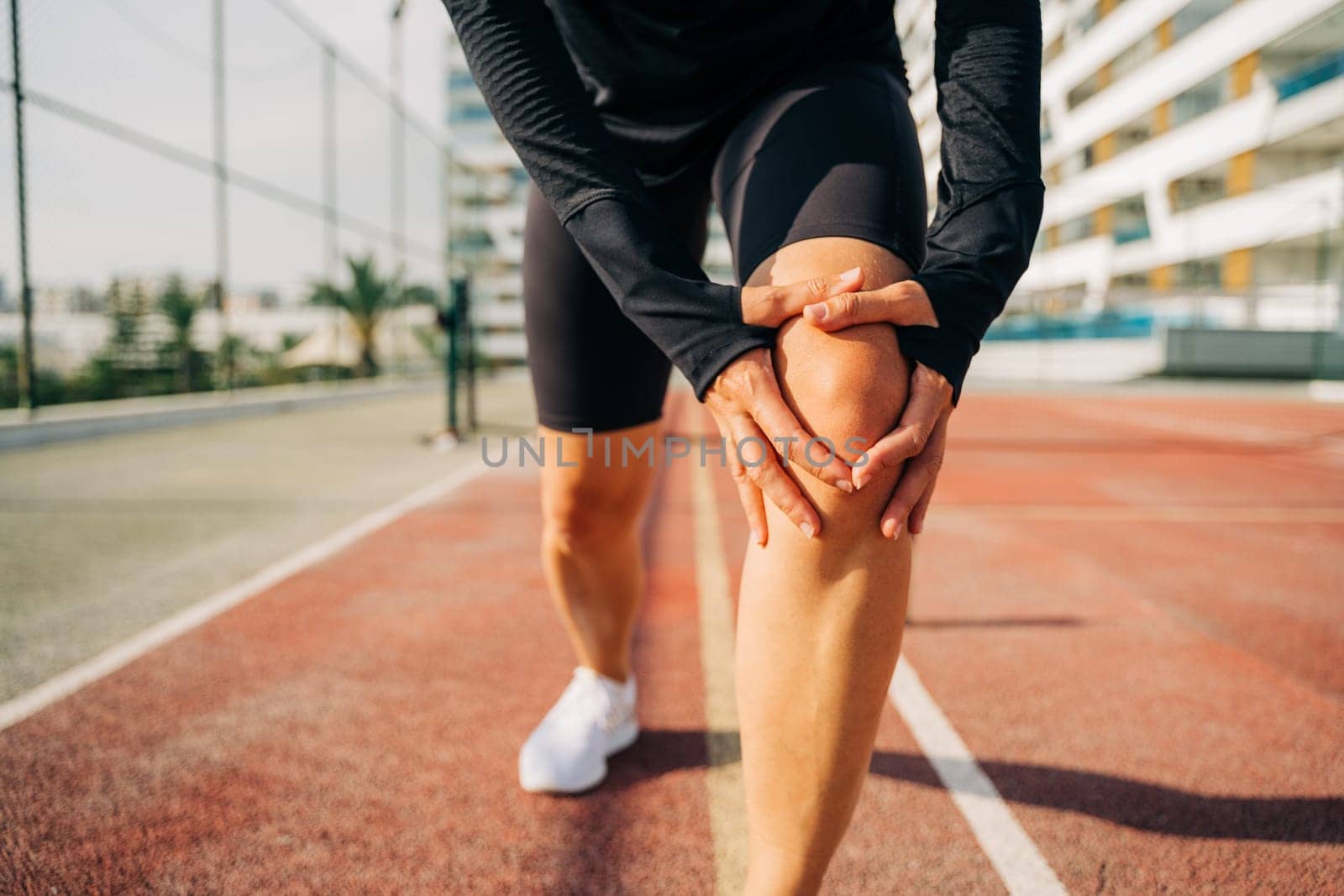 Close up of sport woman holding knee with hands in pain after suffering injury running. Runner woman sit on road with knee injury and pain by Ostanina