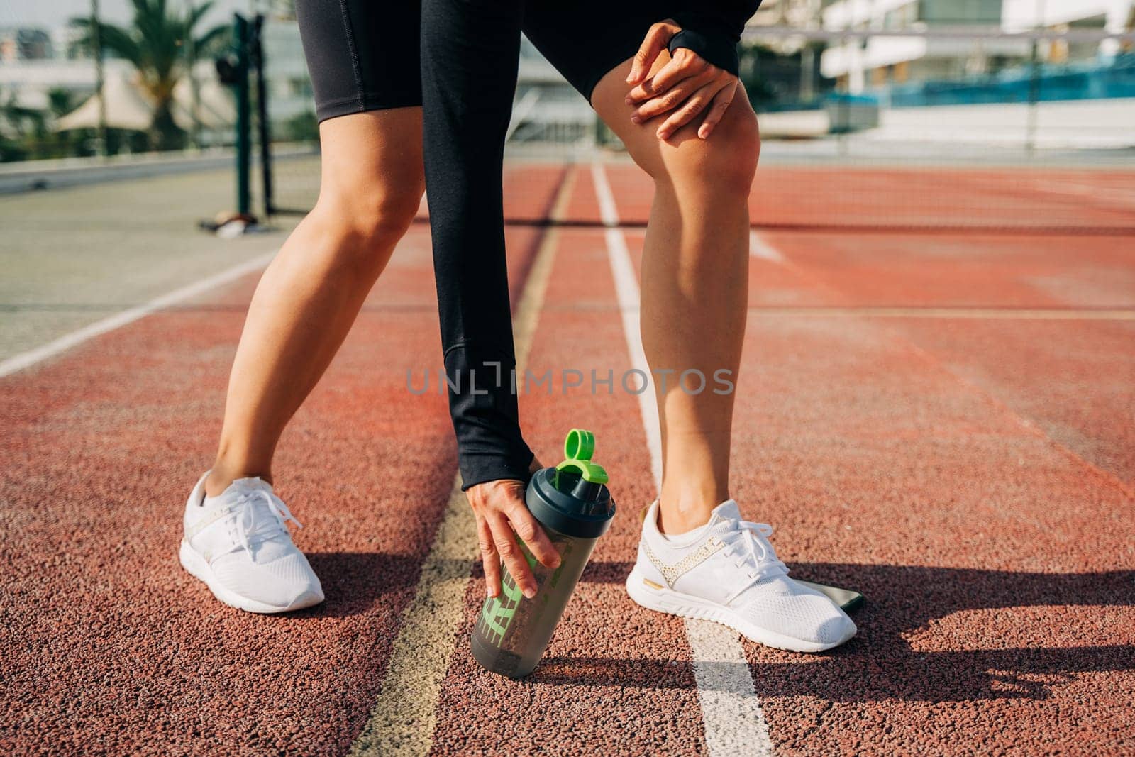Close up of woman runner standing on road and relaxing after sport training. Holding water blender bottle while doing workout in summer city street. by Ostanina