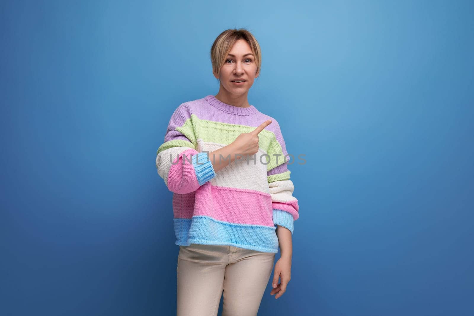 pleasant blond woman in casual outfit shows her hand to the side on a blue background with copy space.