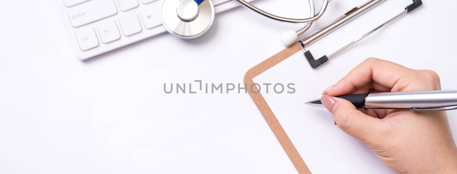 Female doctor writing a medical record case over clipboard on white working table with stethoscope, computer keyboard. Top view, flat lay, copy space by ROMIXIMAGE