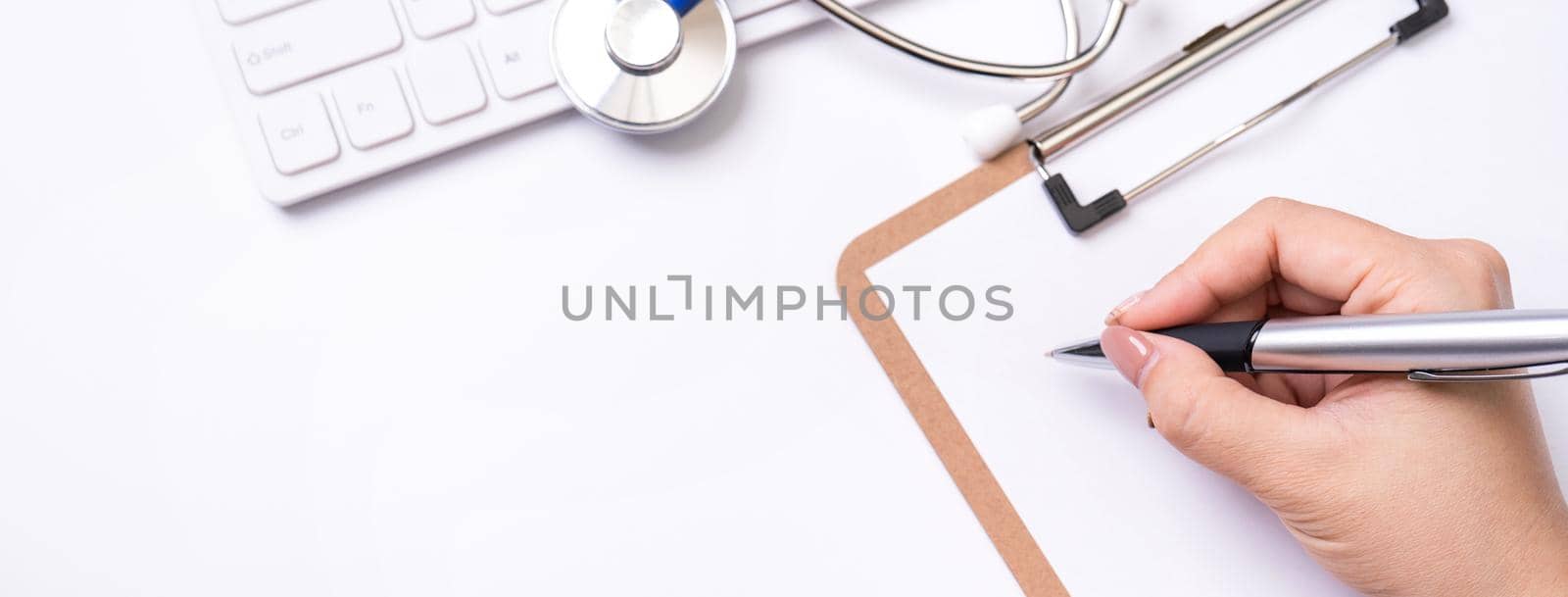 Female doctor writing a medical record case over clipboard on white working table with stethoscope, computer keyboard. Top view, flat lay, copy space