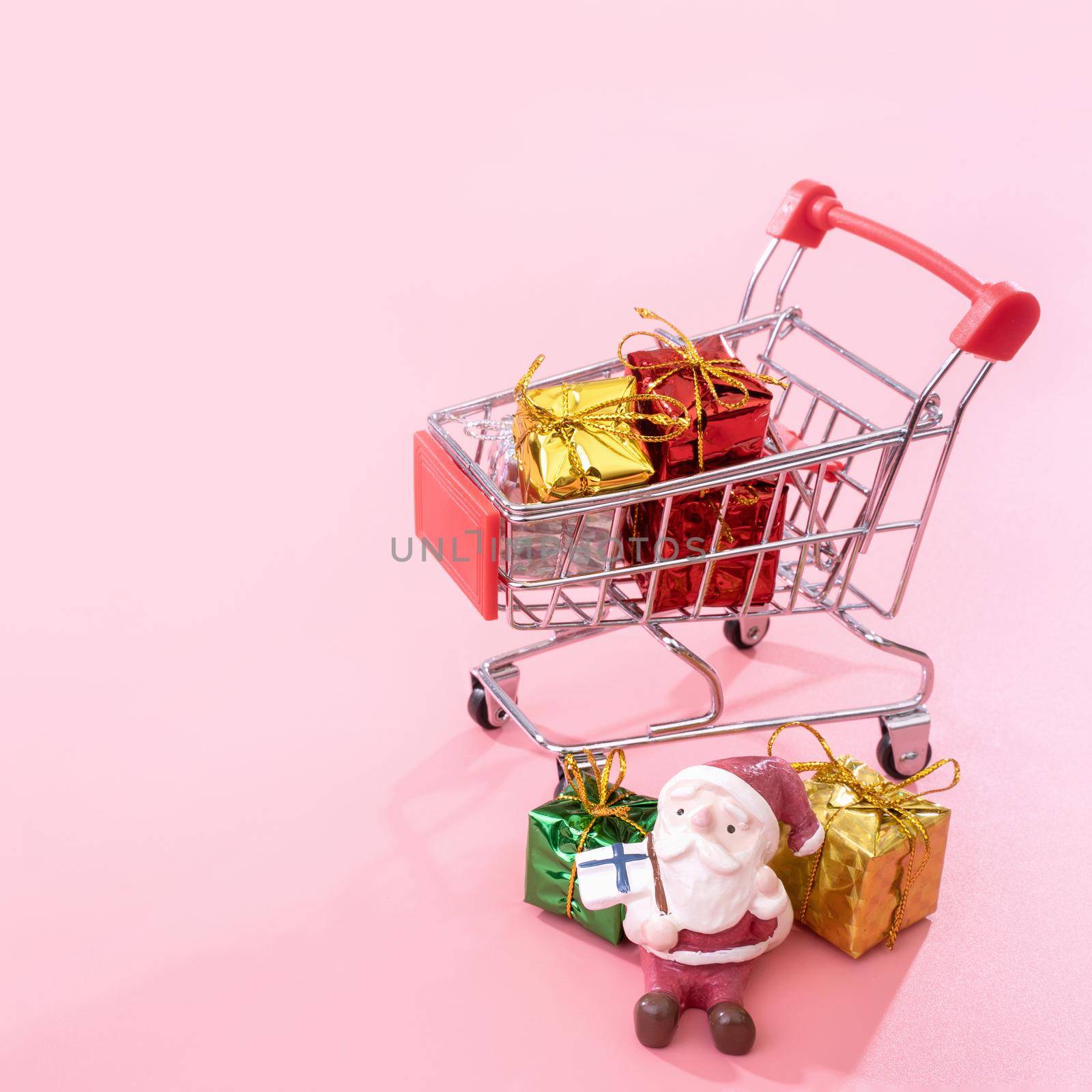 Christmas shopping concept, mini red shop cart trolley with Santa Claus toy and gift box isolated on pale pink background, blank copy space, close up