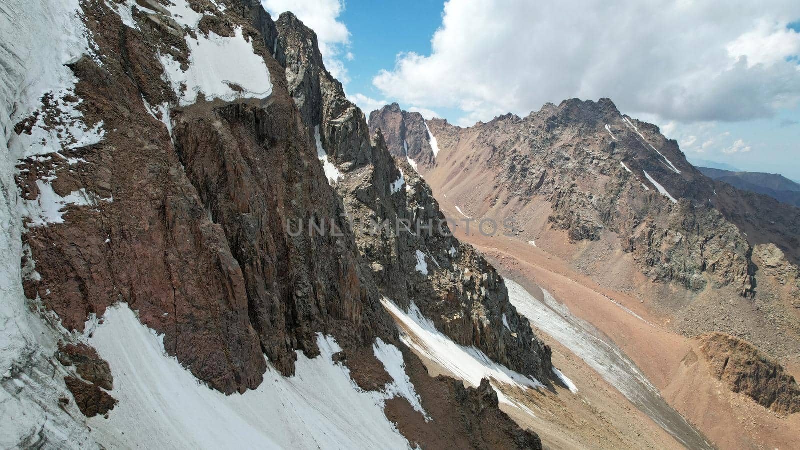 High rocky mountains are sometimes covered with ice and snow. A huge glacier passes between the peaks. The ice is gradually melting. The stones are lying on glacier. Steep cliffs. The view from drone