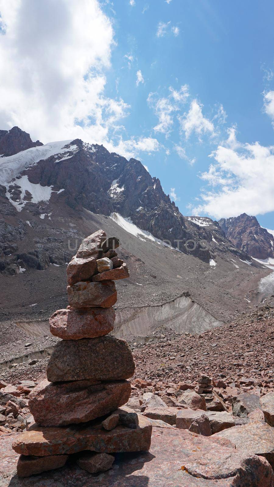 A pyramid of stones as a pointer in the mountains. Big rocks, high peaks and a snowy glacier. A moraine lake can be seen in the distance. The ice is covered with huge rocks. A cairn.