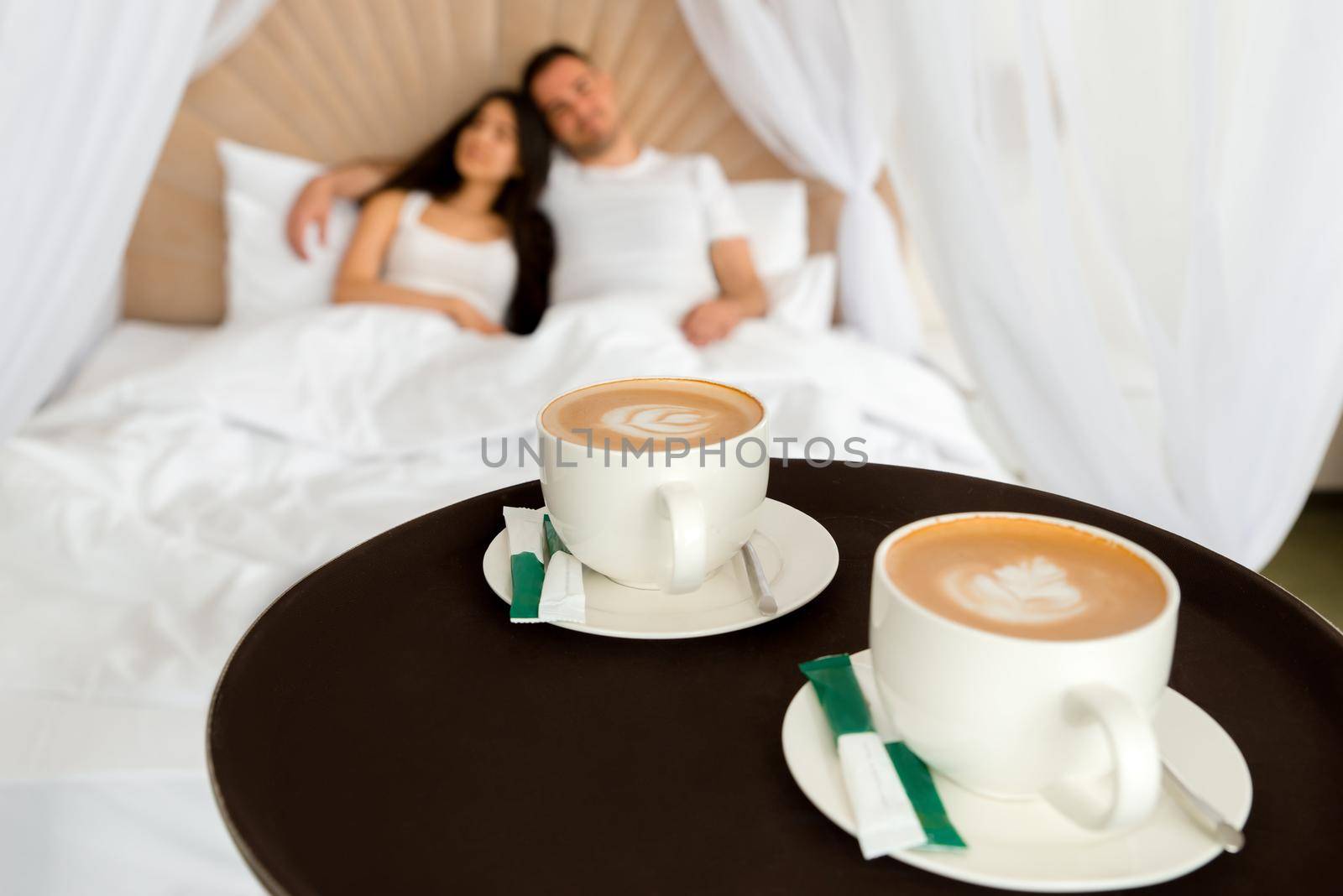 Waiter delivering 2 cups of coffee to a hotel room for young married couple lying in a bed