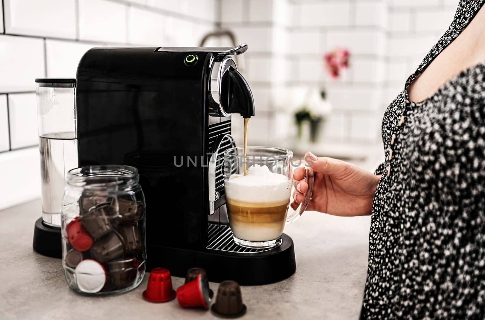 Girl with capsule coffee machine and cappuccino in transparent cup at kitchen. Woman preparing italian caffeine beverage using professional espresso maker
