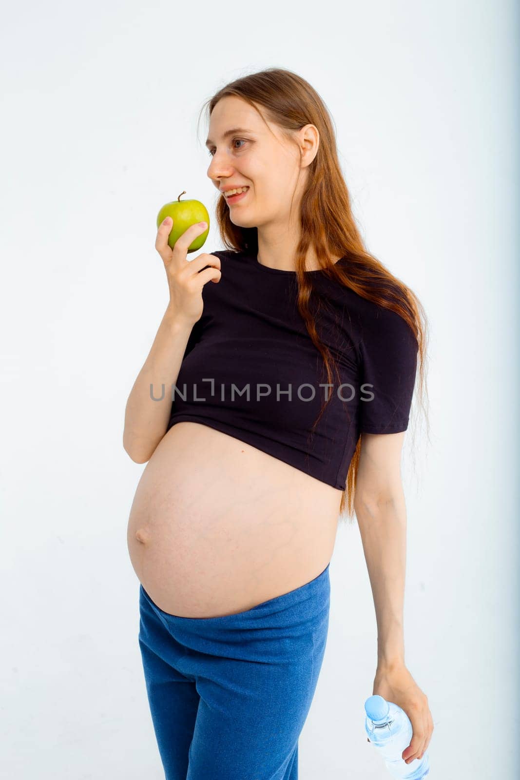 Pregnant woman propose apple for her belly copy space. Beautiful expectant lady having fresh snack, gray studio background. Healthy nutrition and pregnancy concept