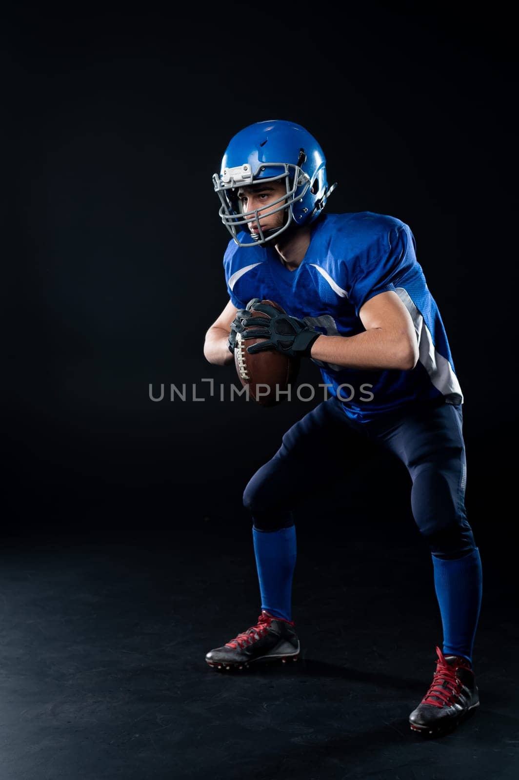 Full length portrait of a man in a blue american football uniform against a black background. Sportsman in a helmet with a ball