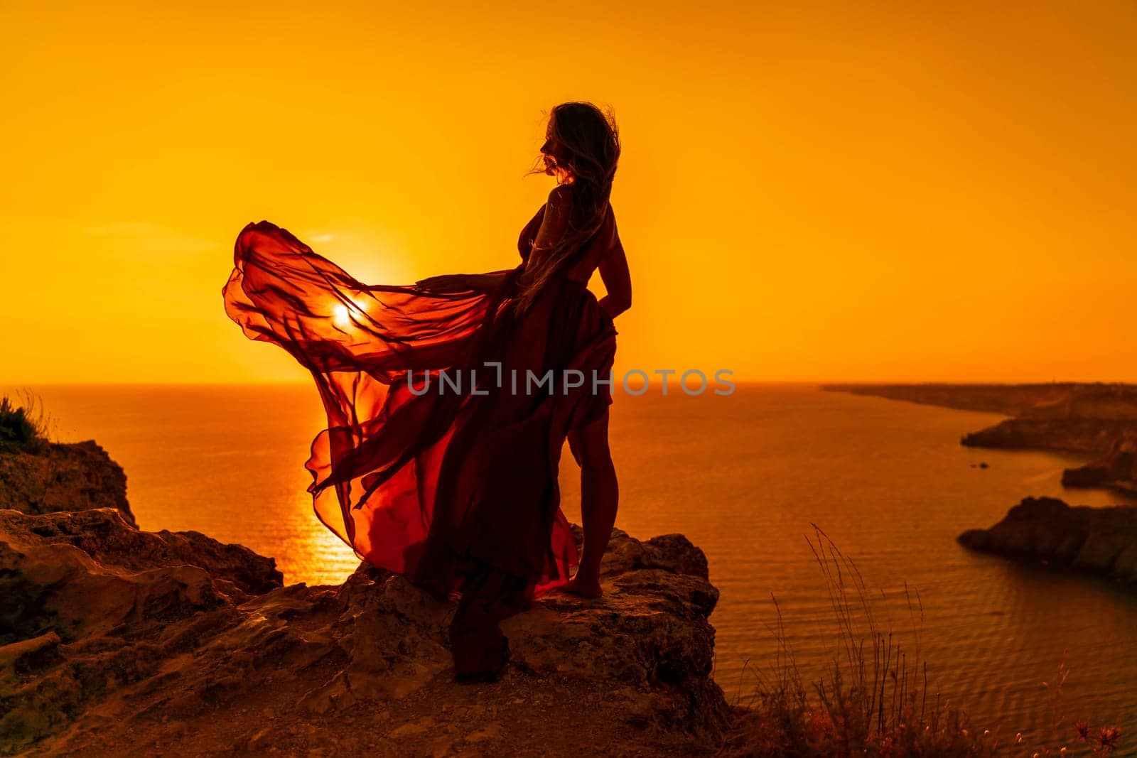 Woman sunset sea red dress, side view a happy beautiful sensual woman in a red long dress posing on a rock high above the sea on sunset. by Matiunina