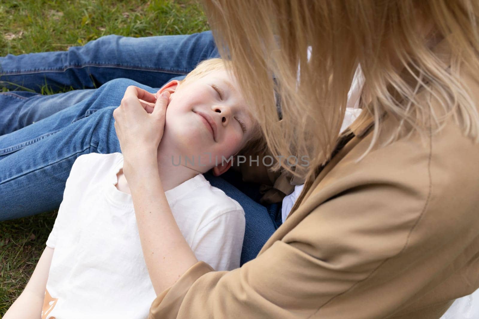 Cute Caucasian 7 yo blonde boy lying on parents knees in park. Mom stroking son's smiling face. Summer time. Parenthood, family leisure time. Love and Care. Closeup Horizontal Plane High quality photo