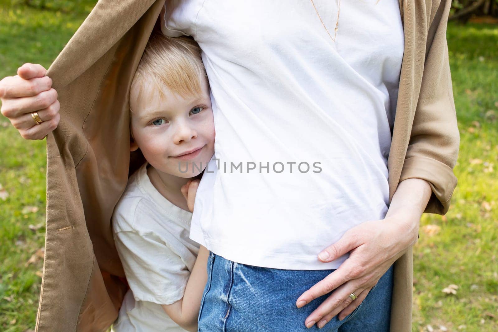 Little Smiling Caucasian 7 Yo Boy Looks Out Mother's Jacket, Hiding Behind Her Back In Park. Mom's Support And Care. Summer Time. Parenthood, Family Leisure Time. Children's Day. Horizontal Plane.