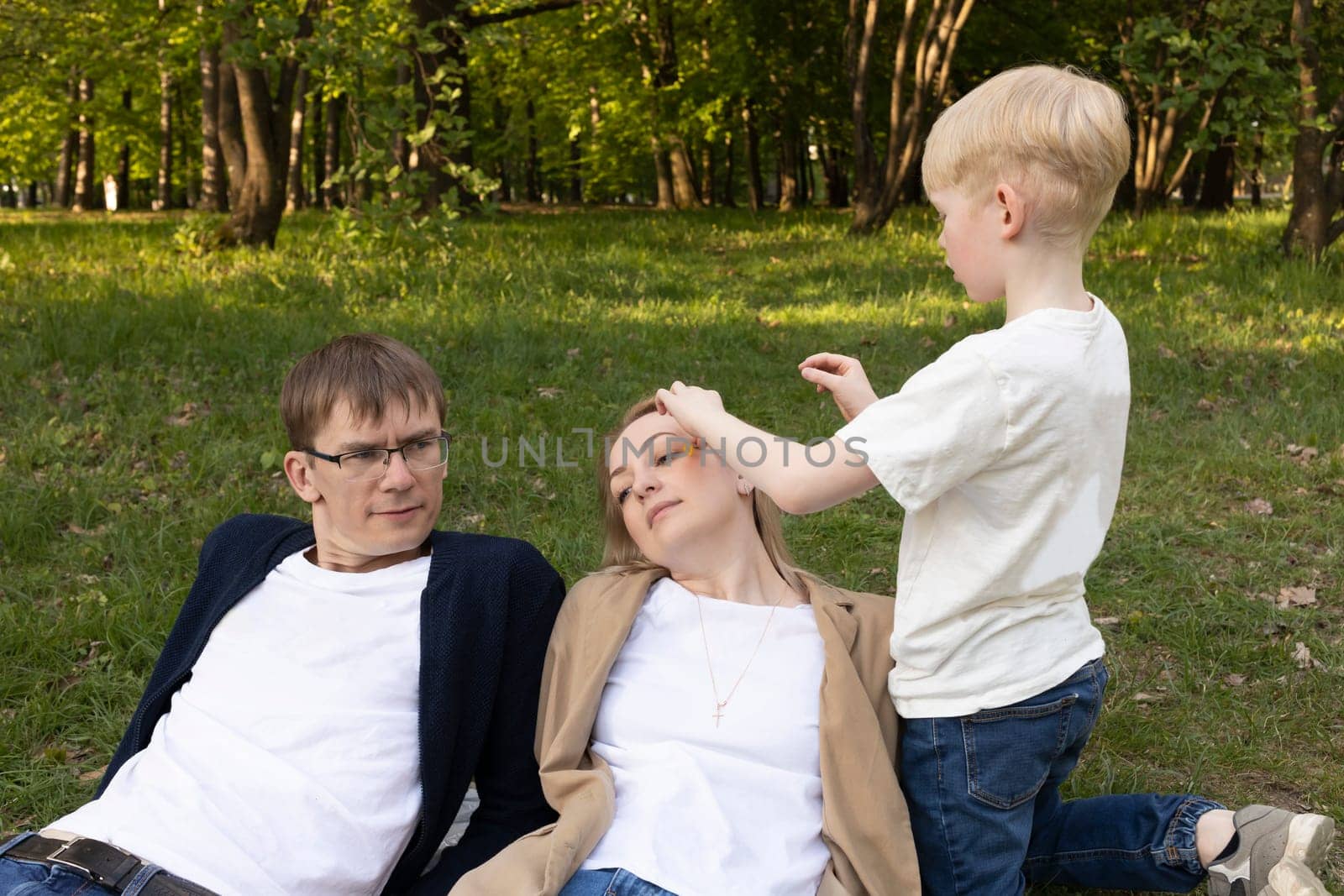 Cute Caucasian 7 Yo Boy Puts, Inserts Flower In Mother's Hair In Park. Mom And Dad Lying On Grass. Summer Time. Parenthood, Family Leisure Time. Love And Care. Horizontal Plane by netatsi