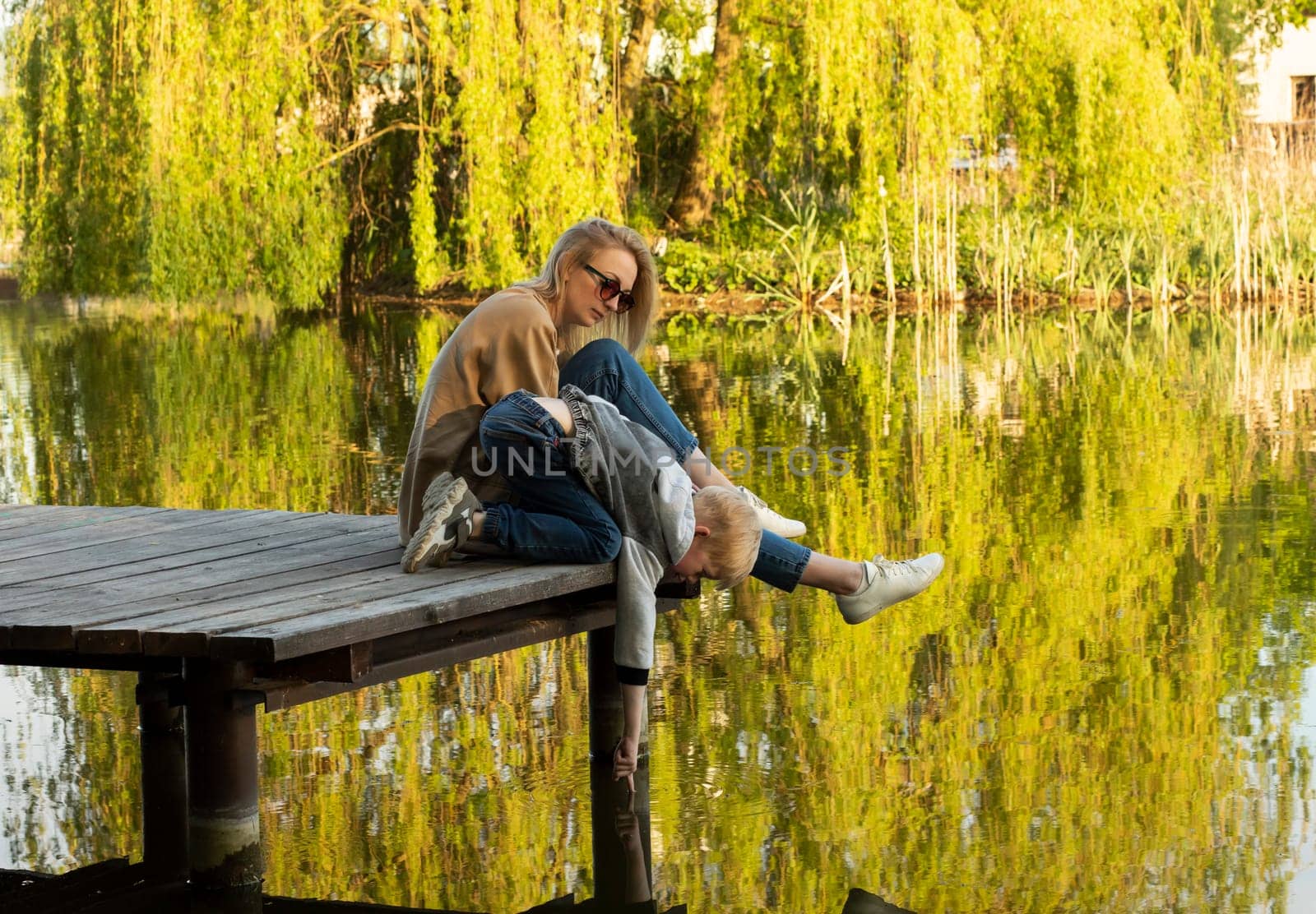 White Mother And Son Sit On Wooden Dock Enjoying Lake View In Summer Time. Child Throws Rocks Into Water. Mom's Support And Care. Motherhood, Family Leisure Time. Children's Day. Horizontal Plane.