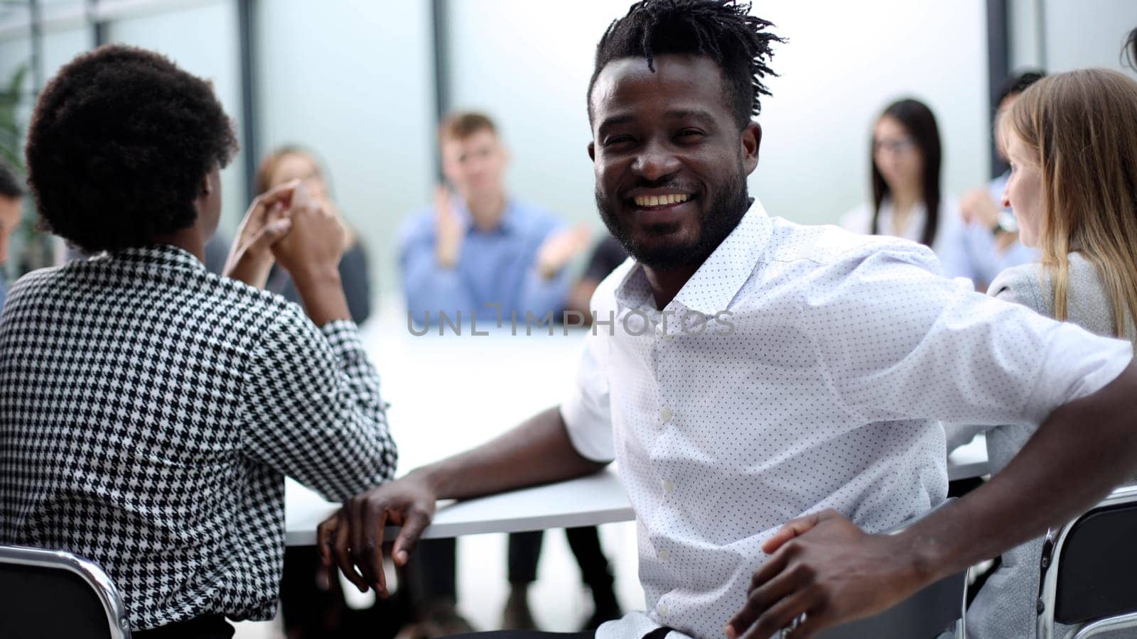 a black man looks at the camera against the background of his colleagues sitting at the table by Prosto