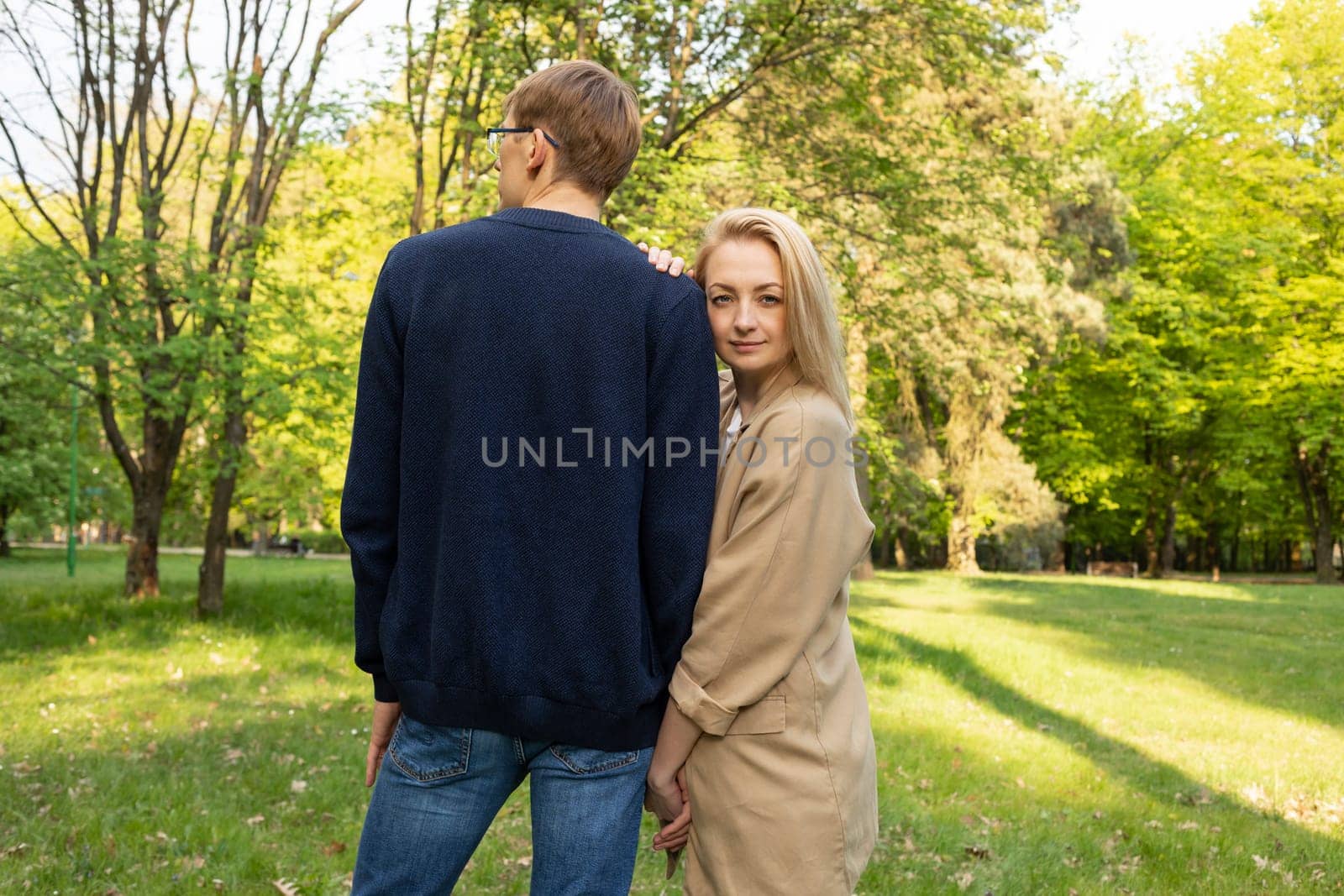 Wife And Husband Walk In Park. Woman Leans Head To Man's Shoulder. Relationship, Togetherness. Enjoying Time Together. Female Looks At Camera, Back View Of Male. Feelings. Horizontal Plane,Summer Time