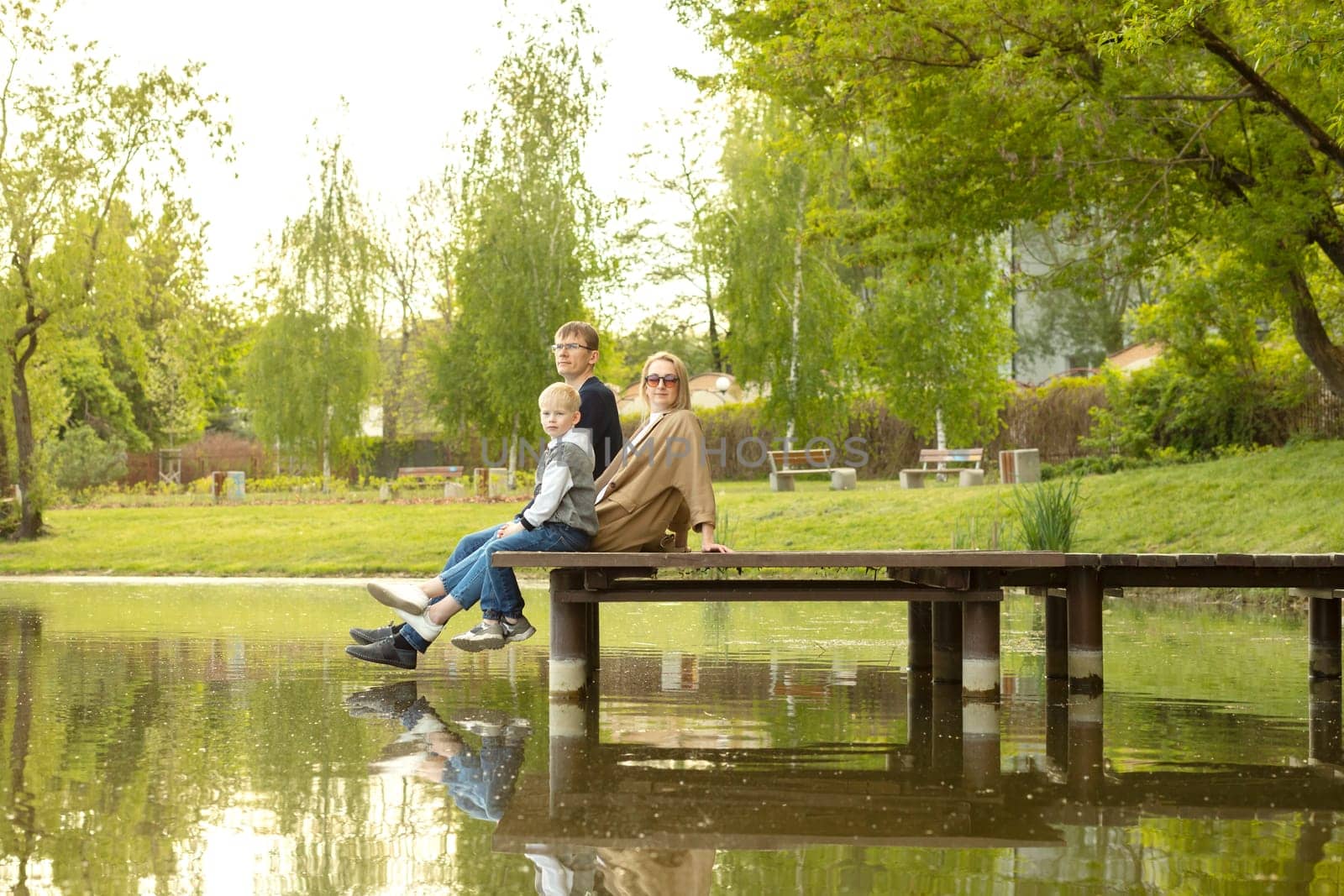Caucasian Family Mother, Father And Son Sit On Wooden Dock Enjoying Lake View In Summer Time. Family Support And Care. Parenthood, Family Leisure Time. Children's Day. Horizontal Plane by netatsi