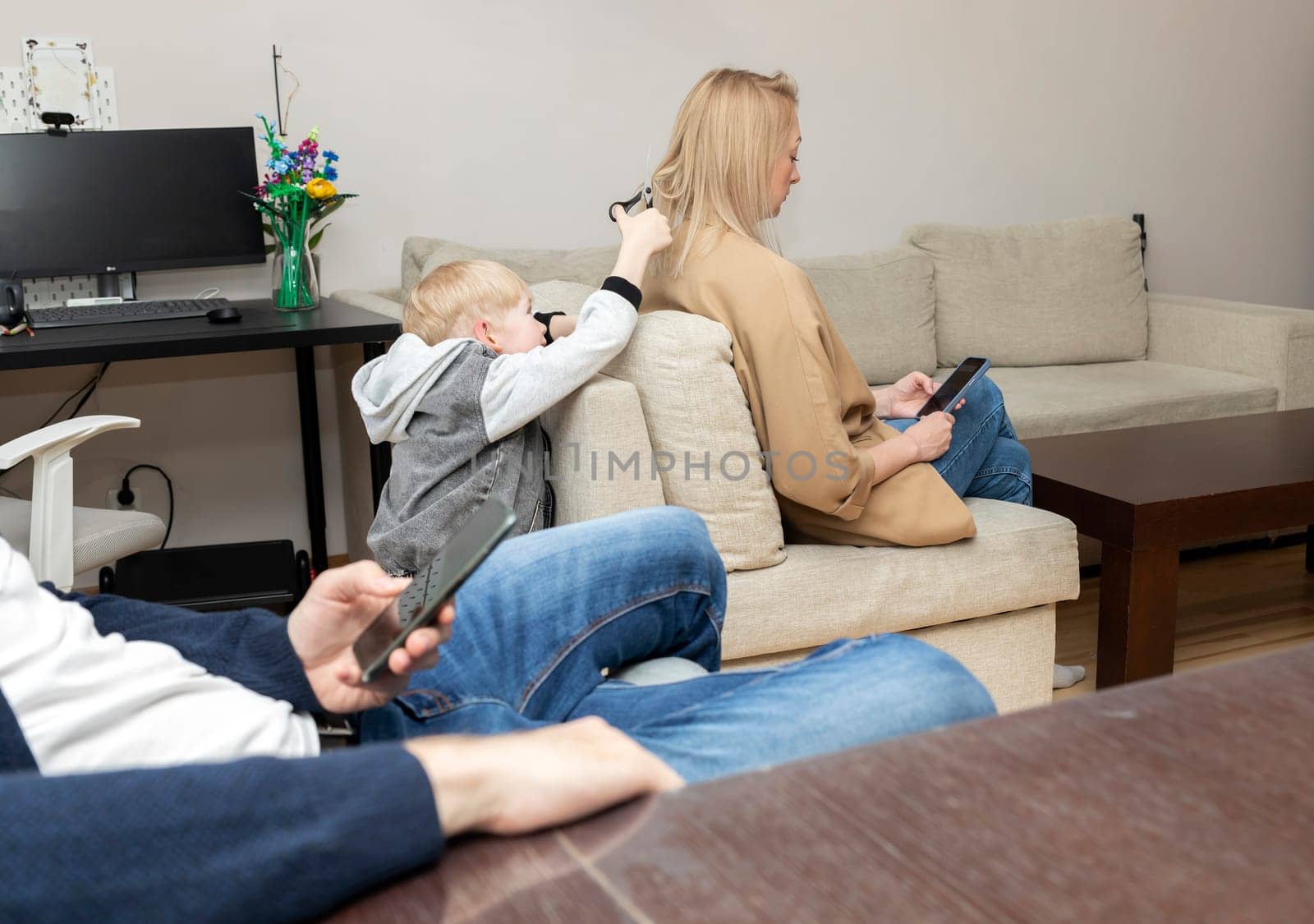 Little Boy Cuts Mother's Hair With Scissors While Parents Look, Sit At Smartphone. Deprivation, Disconnected Family, Emotionally Cold Parents, Social Issue. Horizontal Plane.