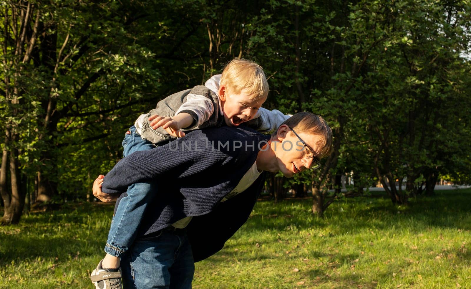 Father Carried Little Happy Child, Son On Back While Walking In Park. Summer Time. Happy Parenthood, Family Leisure Time, Playing Games. Emotional Connection, Love And Care. Horizontal Plane.