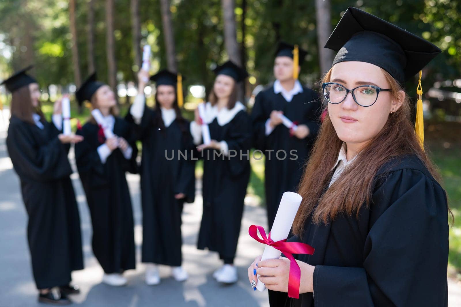 Portrait of a young caucasian woman in glasses and a graduate gown against the background of classmates. A group of graduate students outdoors. by mrwed54