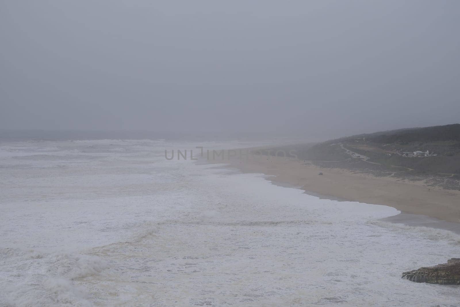 Horizontal photo of gloomy turbulent sea, rough waves, a cape covered with forest is visible in the distance. Foamy waves roll along the sandy shore. Evening time. Twilight sky, Portugal, Nazare