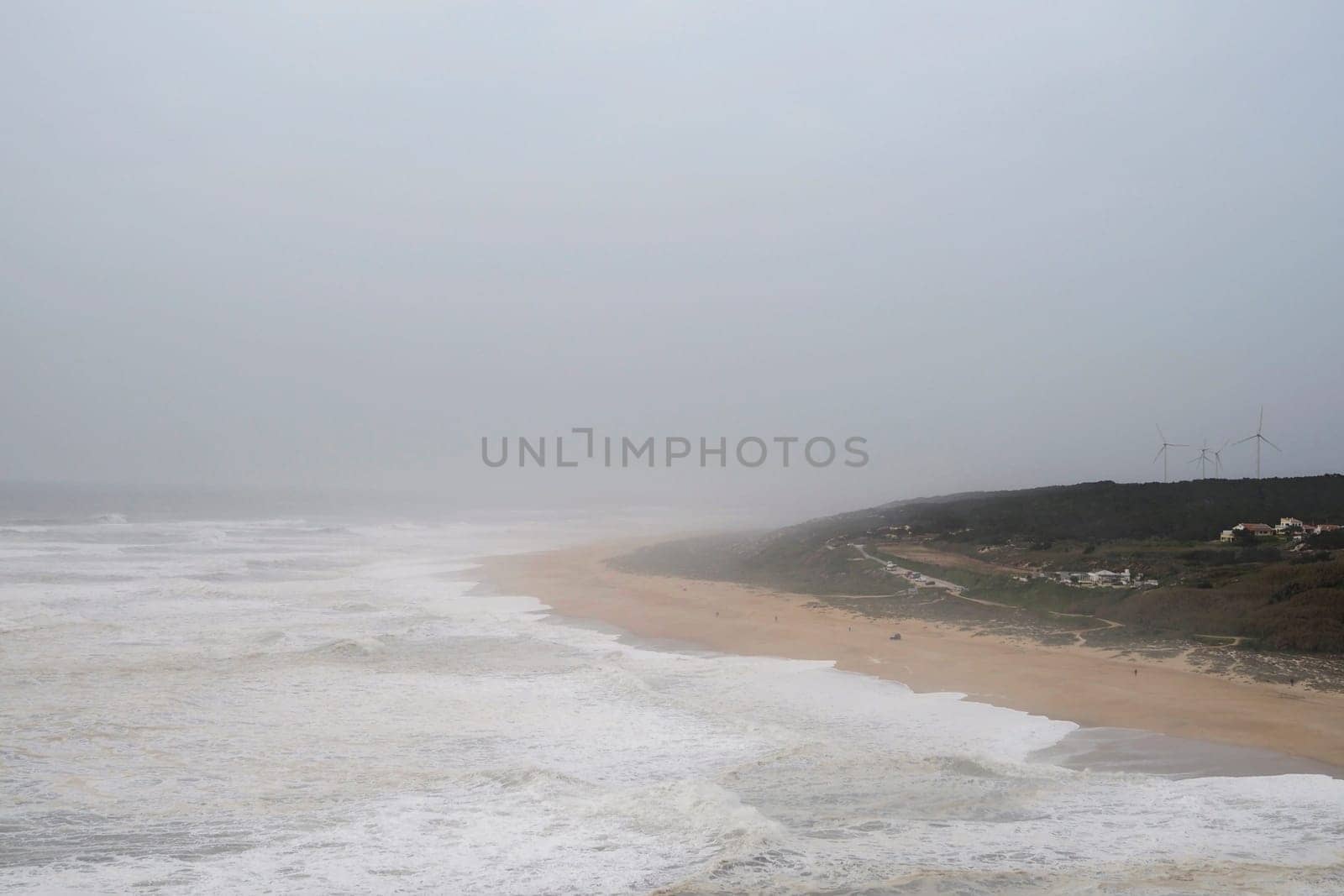 Horizontal photo of overcast stormy sea, turbulent waves, a cape covered with forest is visible in the distance. Windmills and residential houses can be seen in the distance. Foamy waves roll along the sandy shore. Evening time. Twilight sky, Portugal, Nazare