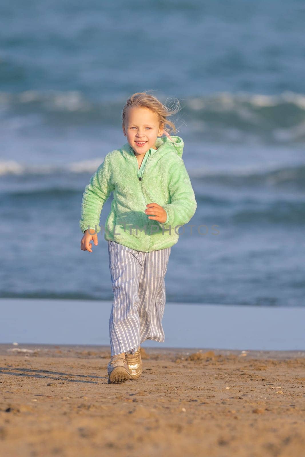 Long shot - vertical photo of girl 5-year-old, a blonde, in a green jacket runs along the beach by the sea, wearing a smile