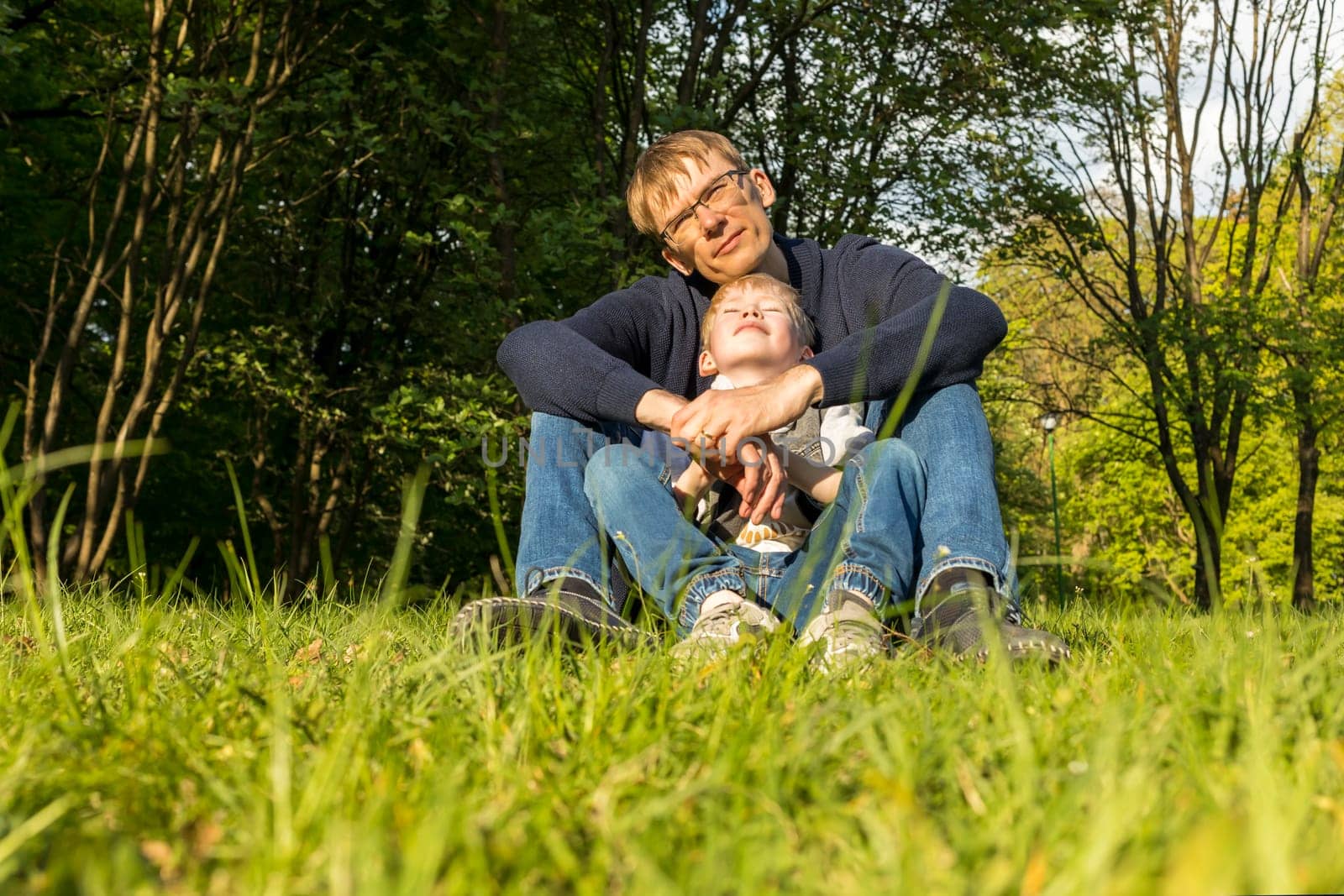 Father And Child, Son Sitting On Grass In Park, Enjoying Time Together. Summer Time. Happy Parenthood, Family Leisure Time, Father's Day. Emotional Connection, Love And Care. Horizontal Plane.