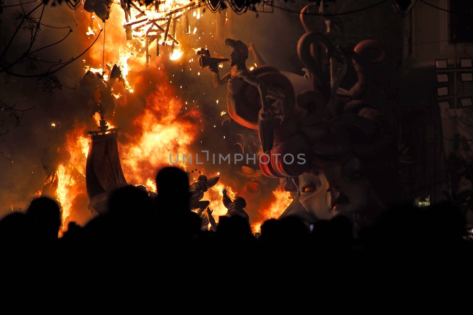 Horizontal photo of people watch Ninote dolls go up in flames in a street lit by lights during the Fallas celebrations in Valencia, Spain