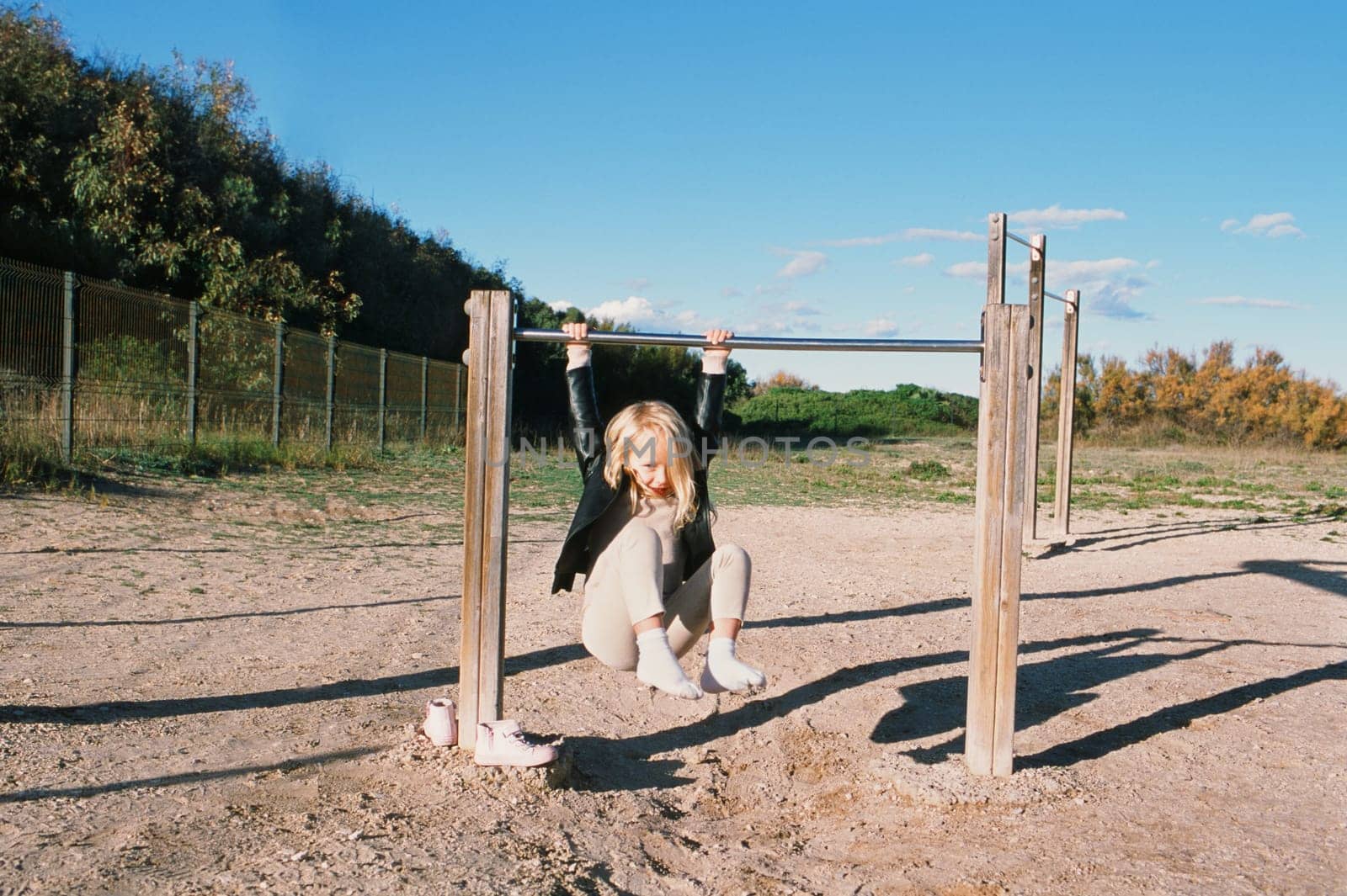 Horizontal photo of a small, 5-year-old blonde girl hangs on a sports bar outside on a playground, surrounded by bushes