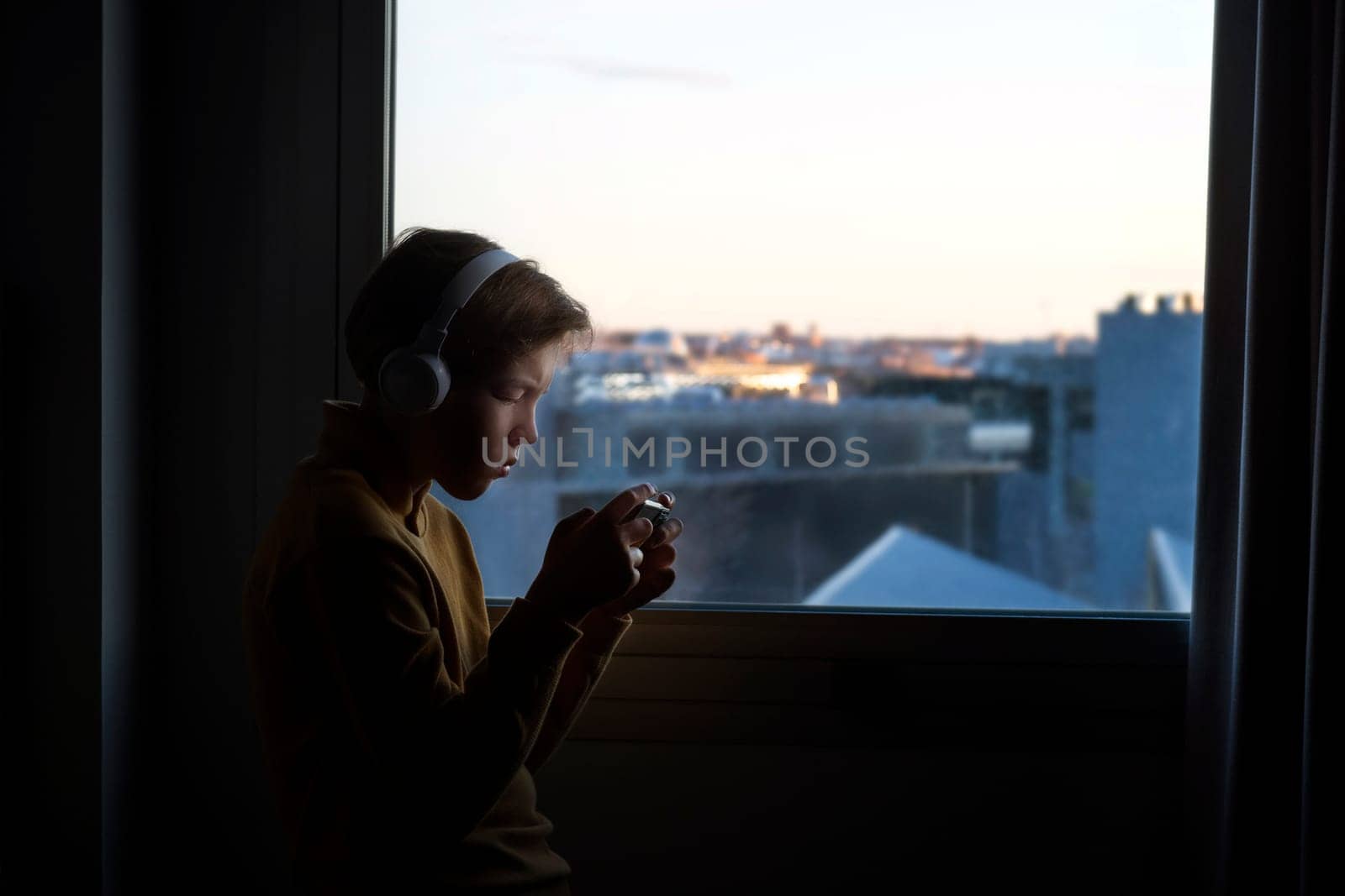 Horizontal photo of a12-year-old caucasian teenager stays in profile at the window against the backdrop of the city, wearing white headphones and playing on his phone