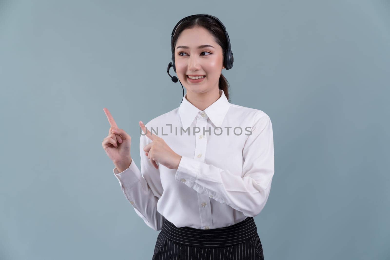 Asian female call center operator with smile face advertises job opportunity, wearing a formal suit and headset pointing finger for product on customizable isolated background. Enthusiastic