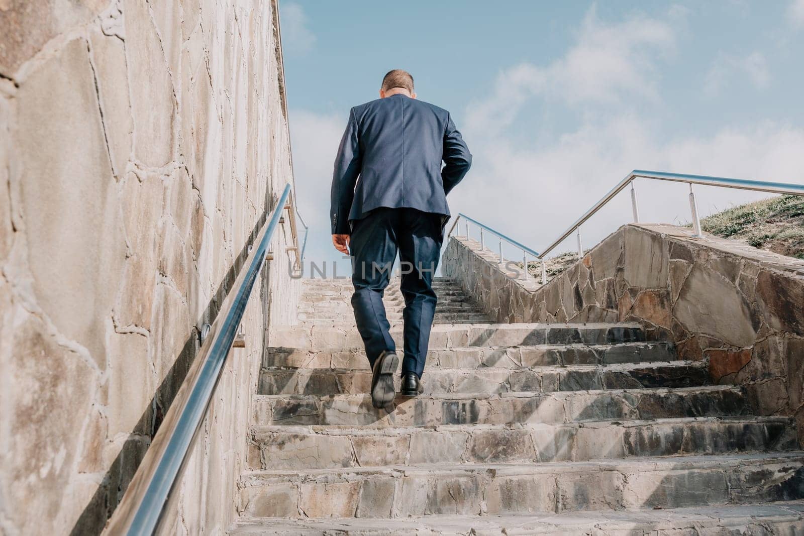 Confident middle age businessman with briefcase walking upstairs. Close-up of businessman wearing blue suit holding bag and coffee cup walking up the stairs by panophotograph