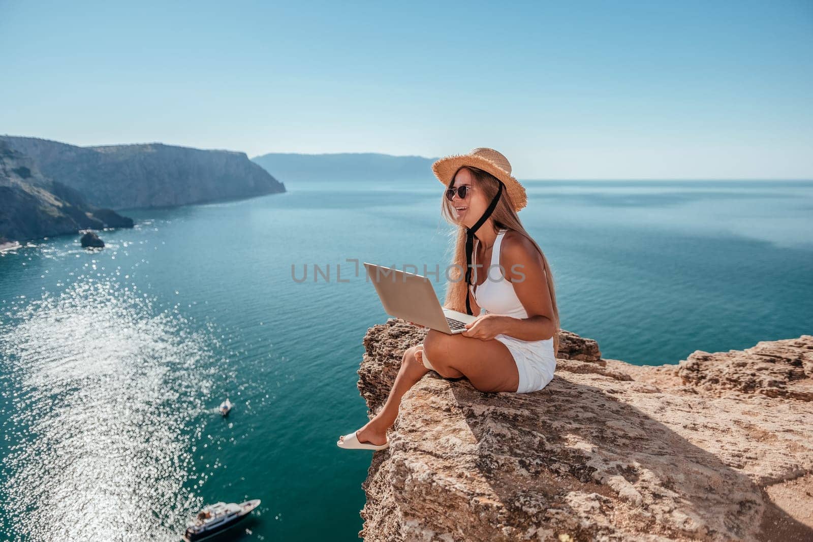 Successful business woman in yellow hat working on laptop by the sea. Pretty lady typing on computer at summer day outdoors. Freelance, travel and holidays concept.